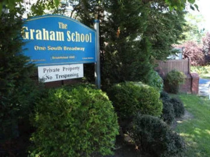 A blue sign displaying "The Graham School," with shrubbery and trees surrounding the sign.