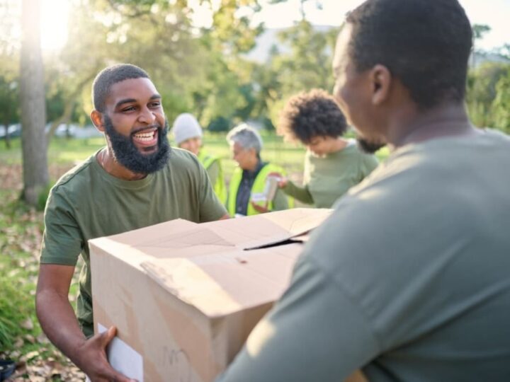 Two men in green t-shirts stand outside in a park. The first man hands the second man a cardboard box filled with fresh produce.